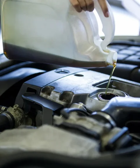 A hand pouring engine oil from a clear container into a car's engine, with engine components visible in the background.