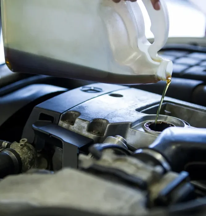 A hand pouring engine oil from a clear container into a car's engine, with engine components visible in the background.