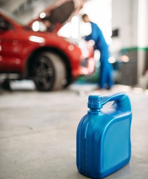 Blue plastic oil container in a garage foreground, with a red car and mechanic in blue overalls working under the opened hood in the blurred background.