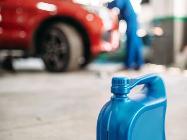Blue plastic oil container in a garage foreground, with a red car and mechanic in blue overalls working under the opened hood in the blurred background.
