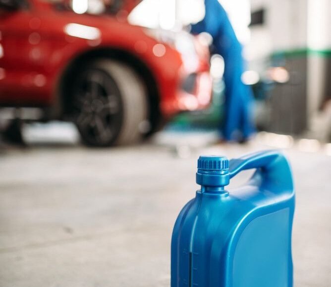 Blue plastic oil container in a garage foreground, with a red car and mechanic in blue overalls working under the opened hood in the blurred background.