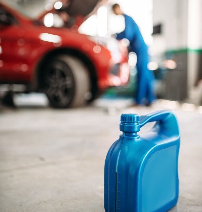 Blue plastic oil container in a garage foreground, with a red car and mechanic in blue overalls working under the opened hood in the blurred background.
