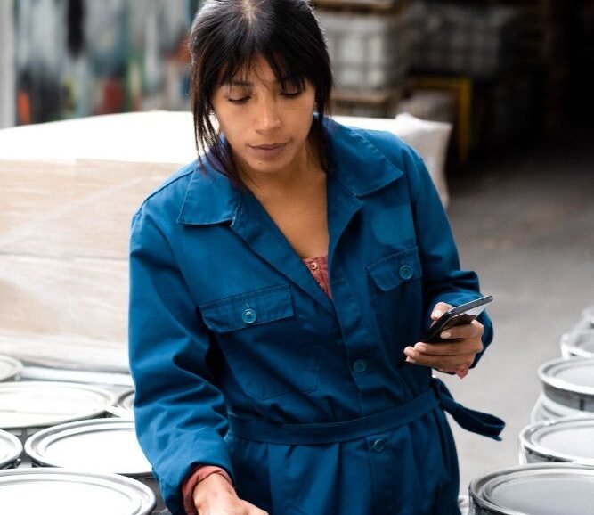 A woman in a blue jumpsuit scans an oil barrel in a warehouse using a handheld device, holding a smartphone. She appears focused and industrious.
