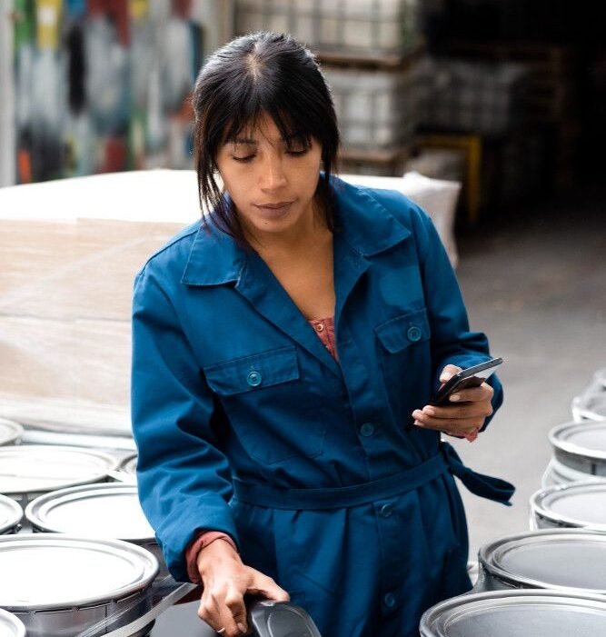 A woman in a blue jumpsuit scans an oil barrel in a warehouse using a handheld device, holding a smartphone. She appears focused and industrious.