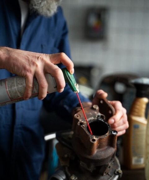 A person in a blue jumpsuit uses a spray lubricant on a rusty metal part in a workshop. The scene conveys focus and a hands-on, repair-oriented atmosphere.
