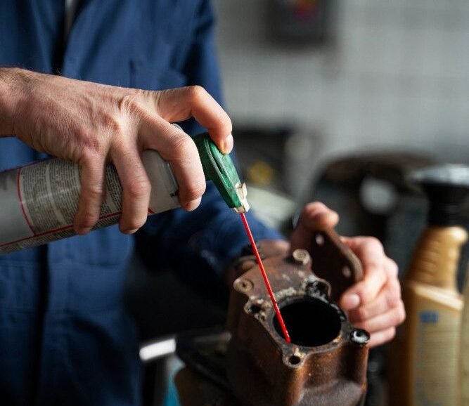 A person in a blue jumpsuit uses a spray lubricant on a rusty metal part in a workshop. The scene conveys focus and a hands-on, repair-oriented atmosphere.
