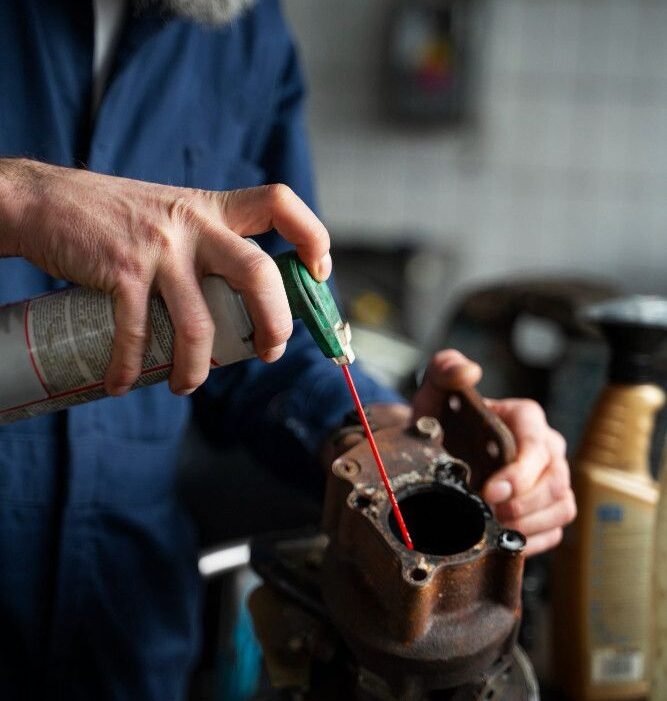 A person in a blue jumpsuit uses a spray lubricant on a rusty metal part in a workshop. The scene conveys focus and a hands-on, repair-oriented atmosphere.