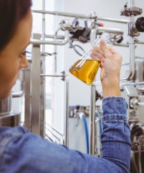 A person in a denim jacket holds a flask containing a yellow liquid, surrounded by industrial equipment in a laboratory setting.