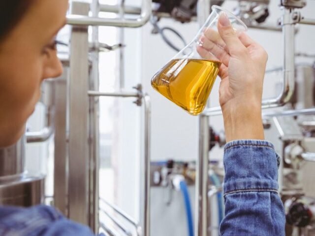 A person in a denim jacket holds a flask containing a yellow liquid, surrounded by industrial equipment in a laboratory setting.