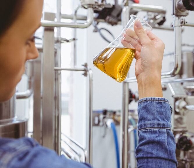 A person in a denim jacket holds a flask containing a yellow liquid, surrounded by industrial equipment in a laboratory setting.