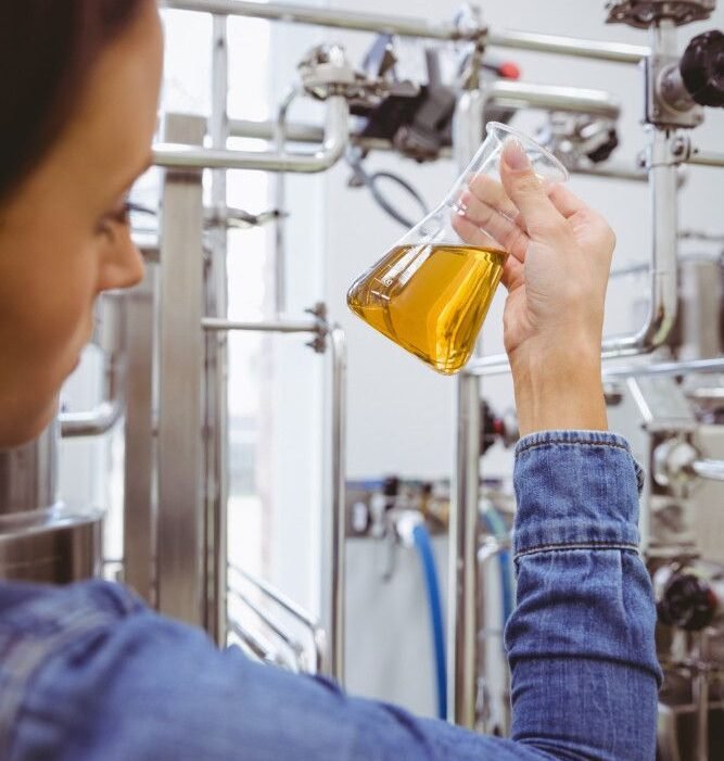 A person in a denim jacket holds a flask containing a yellow liquid, surrounded by industrial equipment in a laboratory setting.